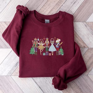 May include: Burgundy crewneck sweatshirt featuring an embroidered design of characters from The Nutcracker ballet. The design includes a nutcracker, a ballerina, and a mouse king, along with Christmas trees and presents. The sweatshirt has a classic fit.