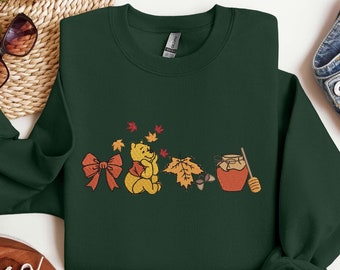 Puede incluir: Una sudadera verde oscuro con un diseño inspirado en Winnie de Pooh que presenta un oso con un lazo rojo, un tarro de miel y hojas de otoño.