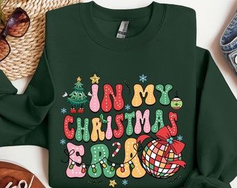 Op de afbeelding: Een donkergroen sweatshirt met een kleurrijk grafisch ontwerp dat "In My Christmas Era" zegt. Het ontwerp bevat een kerstboom, kerstversiering en lampjes.