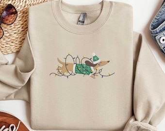 Puede incluir: Sudadera beige de cuello redondo con un perro salchicha bordado que lleva un jersey y un gorro verdes, envuelto en luces navideñas. El perro es marrón y blanco. La sudadera se combina con vaqueros azules y zapatos marrones.
