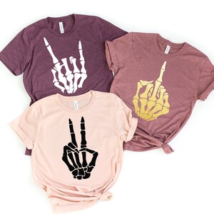 May include: Three t-shirts featuring skeleton hand designs. The first shirt is maroon with a white skeleton hand making a rock and roll sign. The second shirt is maroon with a gold skeleton hand making a middle finger gesture. The third shirt is pink with a black skeleton hand making a peace sign.