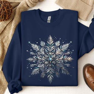 May include: Navy blue sweatshirt featuring a detailed snowflake design in shades of silver, white, and teal. The snowflake is the focal point, with intricate leaf-like patterns and sparkling accents. The sweatshirt is a comfortable, long-sleeved garment.