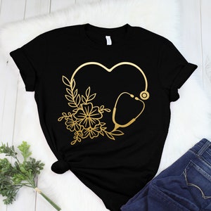 May include: Black t-shirt featuring a gold heart design with a stethoscope and floral accents. The design is centered on the front of the shirt. The shirt is paired with blue jeans on a white fur-like surface.