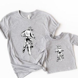May include: Two gray t-shirts with a black and white graphic of a person riding a dirt bike. The larger shirt has a larger graphic of a person riding a dirt bike. The smaller shirt has a smaller graphic of a person riding a dirt bike.