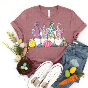 May include: A pink t-shirt with a graphic of four gnomes wearing bunny ears and holding Easter eggs. The gnomes are wearing colorful hats and have white beards. The shirt is paired with blue jeans and white sneakers.