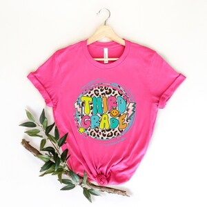 May include: Pink t-shirt with a colorful "Third Grade" graphic on a leopard print background. The graphic includes a smiley face and lightning bolts.