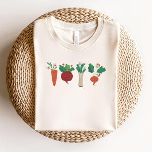 May include: A white t-shirt featuring a graphic of five vegetables, including a carrot, beetroot, onion, and two radishes. The vegetables are illustrated in a cartoon style with green leaves and red, orange, and purple colours.