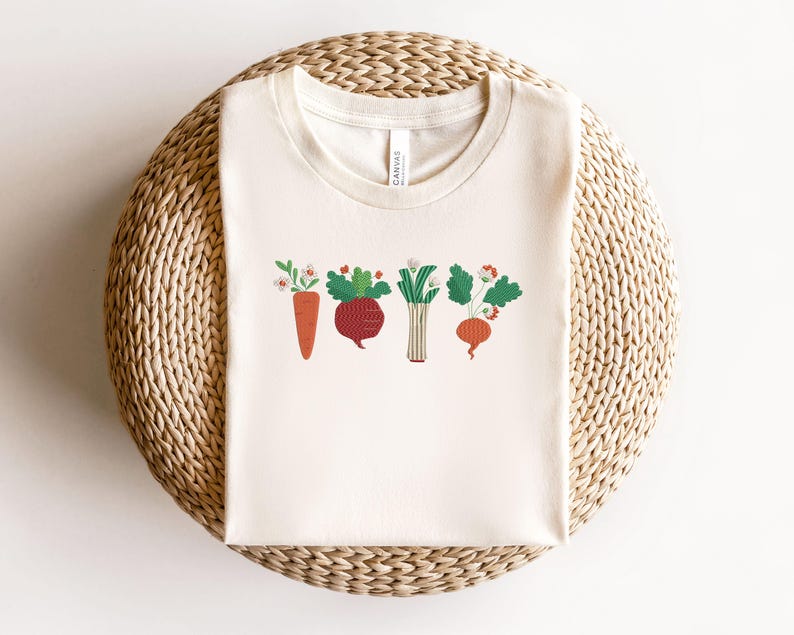 May include: A white t-shirt featuring a graphic of five vegetables, including a carrot, beetroot, onion, and two radishes. The vegetables are illustrated in a cartoon style with green leaves and red, orange, and purple colours.