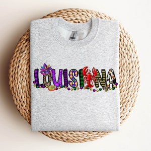May include: A grey sweatshirt featuring the word "LOUISIANA" spelled out in colourful letters. Each letter is decorated with Mardi Gras themed imagery, including masks, beads, and a crawfish.