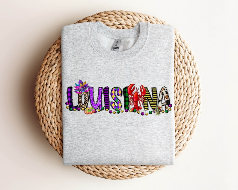 May include: A grey sweatshirt featuring the word "LOUISIANA" spelled out in colourful letters. Each letter is decorated with Mardi Gras themed imagery, including masks, beads, and a crawfish.