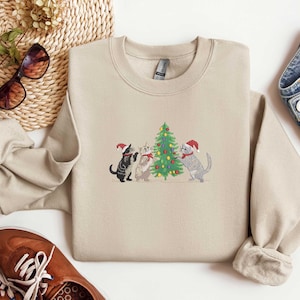 K&ouml;nnte beinhalten: Beiges Sweatshirt mit Rundhalsausschnitt und gesticktem Design von drei Katzen mit Weihnachtsm&uuml;tzen um einen geschm&uuml;ckten Weihnachtsbaum. Die Katzen sind schwarz, grau und wei&szlig;. Das Sweatshirt wird mit blauen Jeans und braunen Schuhen kombiniert.