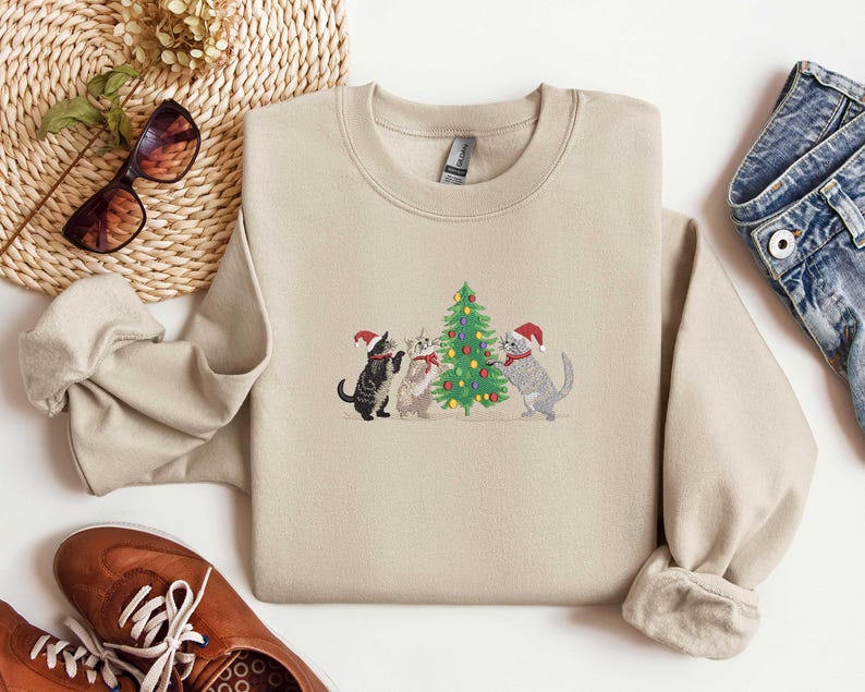 K&ouml;nnte beinhalten: Beiges Sweatshirt mit Rundhalsausschnitt und gesticktem Design von drei Katzen mit Weihnachtsm&uuml;tzen um einen geschm&uuml;ckten Weihnachtsbaum. Die Katzen sind schwarz, grau und wei&szlig;. Das Sweatshirt wird mit blauen Jeans und braunen Schuhen kombiniert.