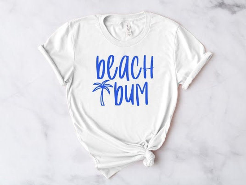 以下が含まれることがあります： ビーチバームと青いヤシの木のグラフィックが書かれた白いTシャツです。