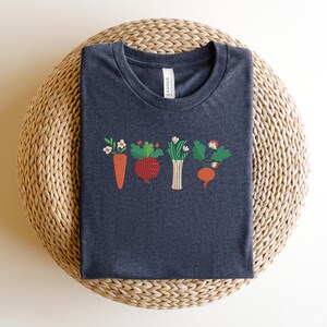 May include: A dark grey t-shirt with an embroidered design of a carrot, beetroot, onion, and radish with green leaves and white flowers.