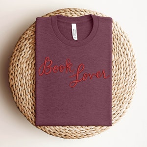 K&ouml;nnte beinhalten: Ein maroonfarbenes T-Shirt mit den Worten "Book Lover" in roter Stickerei.