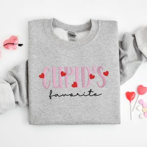 May include: A gray sweatshirt with the text "Cupid's favorite" in pink lettering with red hearts.