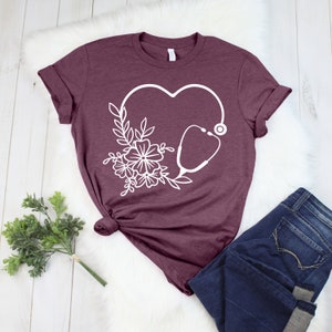 May include: Burgundy t-shirt featuring a white heart-shaped design with a stethoscope and floral elements. The shirt is paired with dark blue jeans. The design is a medical or healthcare theme.