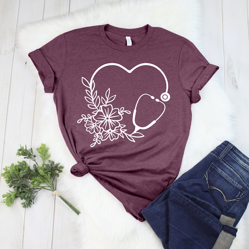 May include: Burgundy t-shirt featuring a white heart-shaped design with a stethoscope and floral elements. The shirt is paired with dark blue jeans. The design is a medical or healthcare theme.