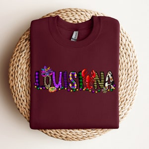 May include: Burgundy t-shirt featuring a colourful graphic design that reads "LOUISIANA" with Mardi Gras beads, a mask, a crawfish, and a fleur-de-lis.