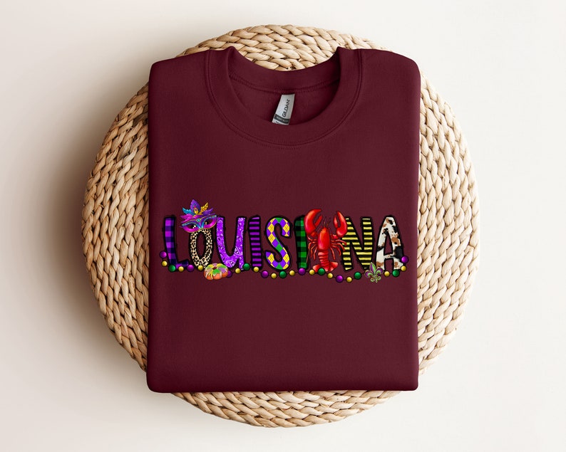 May include: Burgundy t-shirt featuring a colourful graphic design that reads "LOUISIANA" with Mardi Gras beads, a mask, a crawfish, and a fleur-de-lis.