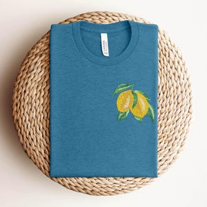 Puede incluir: Una camiseta azul turquesa con un dise&ntilde;o de lim&oacute;n bordado. El lim&oacute;n es amarillo con hojas verdes.