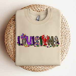 May include: A tan sweatshirt with the word "LOUISIANA" spelled out in colourful letters with Mardi Gras themed decorations.