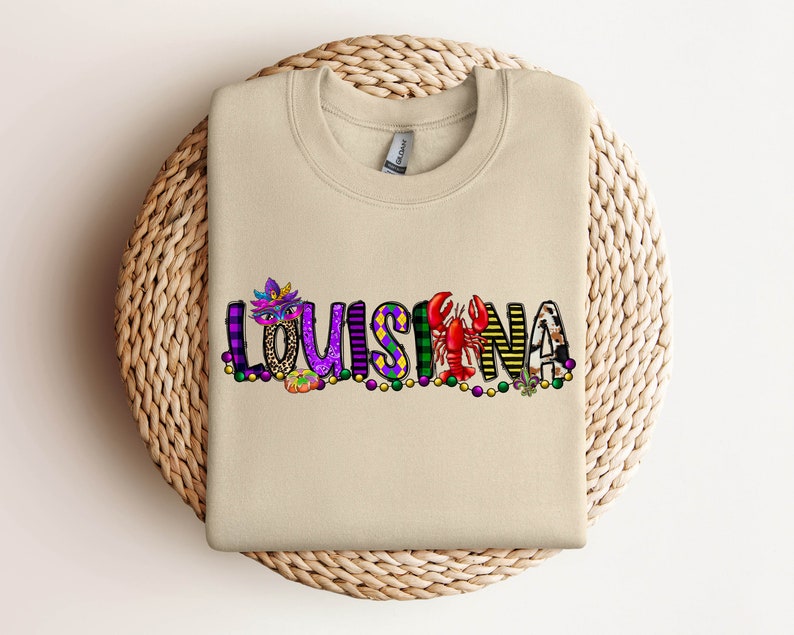 May include: A tan sweatshirt with the word "LOUISIANA" spelled out in colourful letters with Mardi Gras themed decorations.