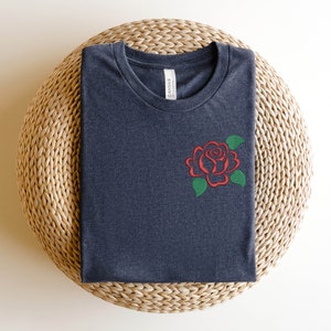 May include: A dark gray t-shirt with a red rose embroidered on the left chest. The rose has green leaves and is a simple design.