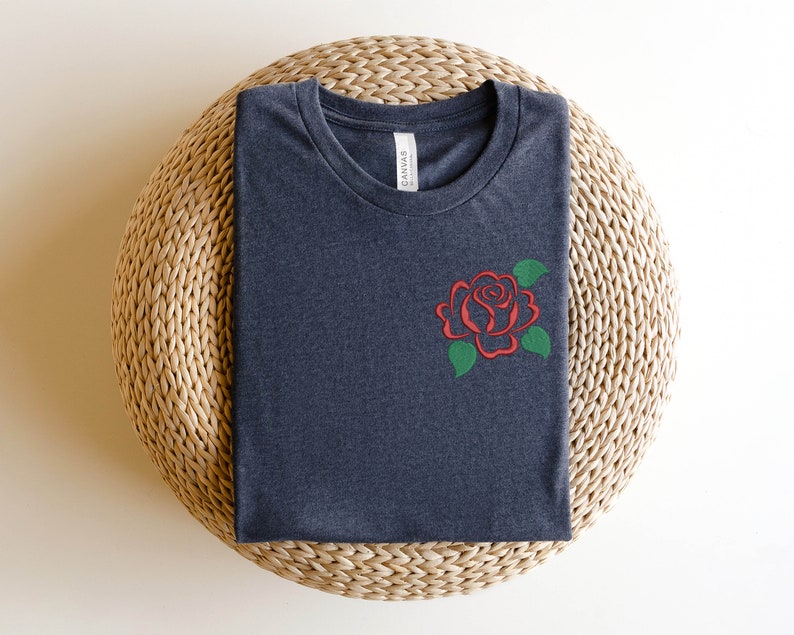 May include: A dark gray t-shirt with a red rose embroidered on the left chest. The rose has green leaves and is a simple design.
