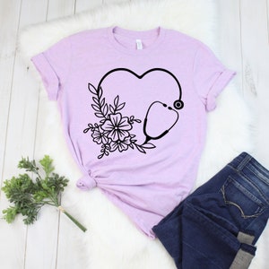 May include: A light purple t-shirt featuring a black heart-shaped design with a stethoscope and floral accents. The shirt is paired with dark blue jeans. The design is a medical or healthcare theme.