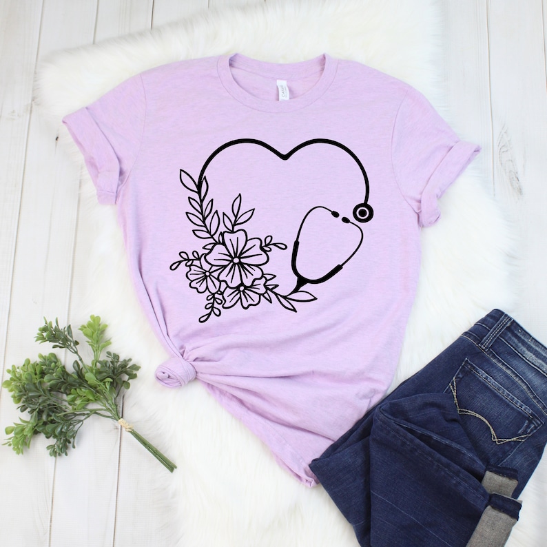 May include: A light purple t-shirt featuring a black heart-shaped design with a stethoscope and floral accents. The shirt is paired with dark blue jeans. The design is a medical or healthcare theme.