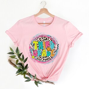 May include: Pink t-shirt with a colorful "Third Grade" graphic on a leopard print background.