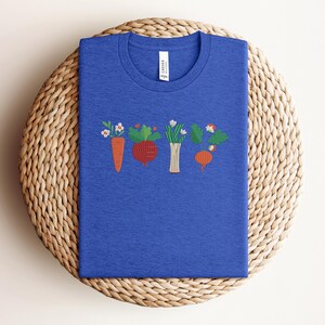 May include: A royal blue t-shirt featuring an embroidered design of a row of vegetables, including a carrot, a beetroot, a bunch of spring onions, and a turnip.