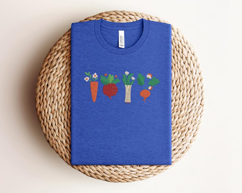 May include: A royal blue t-shirt featuring an embroidered design of a row of vegetables, including a carrot, a beetroot, a bunch of spring onions, and a turnip.