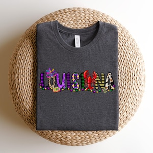 May include: A dark grey t-shirt with the word "LOUISIANA" spelled out in colourful letters. Each letter is decorated with Mardi Gras themed designs, including beads, masks, and a crawfish.
