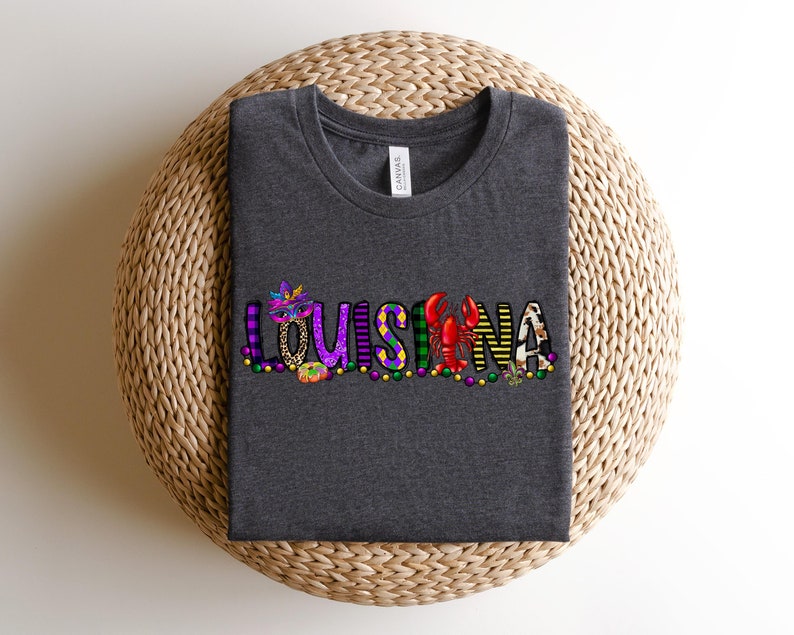 May include: A dark grey t-shirt with the word "LOUISIANA" spelled out in colourful letters. Each letter is decorated with Mardi Gras themed designs, including beads, masks, and a crawfish.