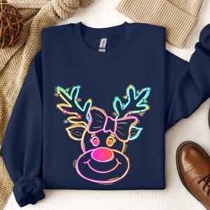 May include: Navy blue sweatshirt featuring a neon reindeer design. The reindeer has antlers adorned with colorful lights, a pink nose, and a bow. The sweatshirt is a crew neck style, perfect for the holiday season.