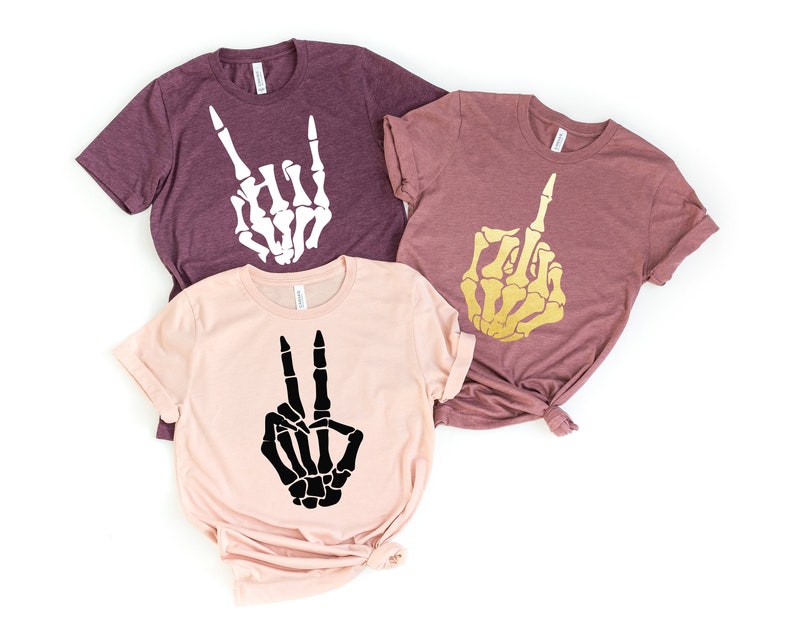 May include: Three t-shirts featuring skeleton hand designs. The first shirt is maroon with a white skeleton hand making a rock and roll sign. The second shirt is maroon with a gold skeleton hand making a middle finger gesture. The third shirt is pink with a black skeleton hand making a peace sign.