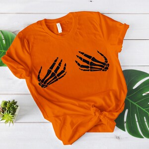 Puede incluir: Camiseta naranja con un gr&aacute;fico de mano de esqueleto negro en la parte delantera. La camiseta est&aacute; doblada y colocada sobre una superficie de madera blanca.
