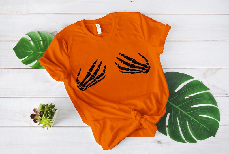 Puede incluir: Camiseta naranja con un gr&aacute;fico de mano de esqueleto negro en la parte delantera. La camiseta est&aacute; doblada y colocada sobre una superficie de madera blanca.
