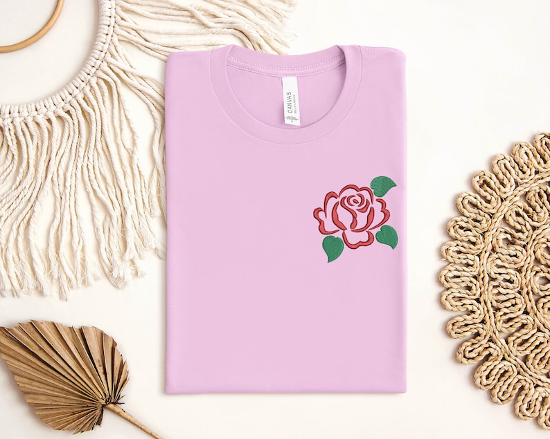 May include: A light pink t-shirt with a red rose embroidered on the front. The rose has green leaves.