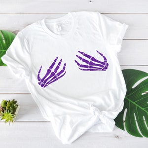 Puede incluir: Camiseta blanca con un dise&ntilde;o de mano de esqueleto morado en la parte delantera. El dise&ntilde;o es de dos manos con los dedos extendidos.