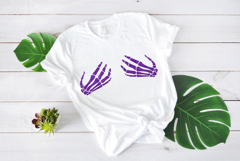 Puede incluir: Camiseta blanca con un dise&ntilde;o de mano de esqueleto morado en la parte delantera. El dise&ntilde;o es de dos manos con los dedos extendidos.