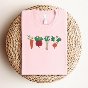 May include: Pink t-shirt featuring a graphic of a row of colourful vegetables, including a carrot, beetroot, leek, and turnip.