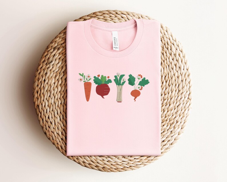 May include: Pink t-shirt featuring a graphic of a row of colourful vegetables, including a carrot, beetroot, leek, and turnip.
