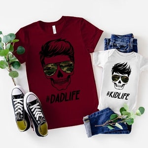 May include: A red t-shirt with a skull graphic wearing sunglasses and the text "#DADLIFE". A white onesie with a skull graphic wearing sunglasses and the text "#KIDLIFE".