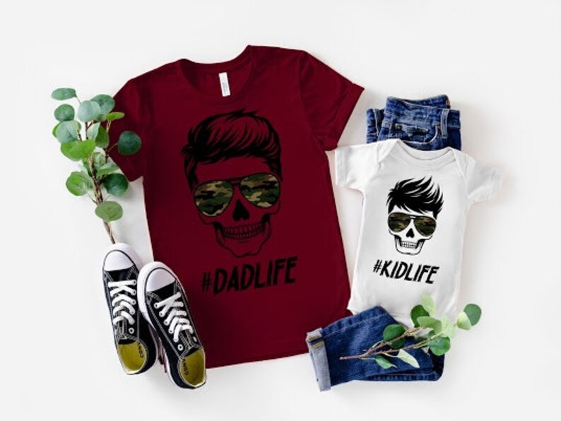 May include: A red t-shirt with a skull graphic wearing sunglasses and the text "#DADLIFE". A white onesie with a skull graphic wearing sunglasses and the text "#KIDLIFE".