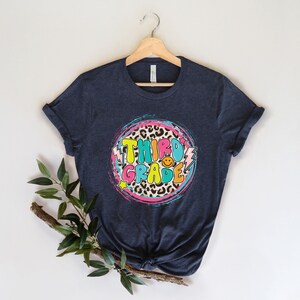 May include: A dark blue t-shirt with a colorful graphic design that says "Third Grade" in a retro font. The graphic features a leopard print circle with pink, blue, and yellow accents.