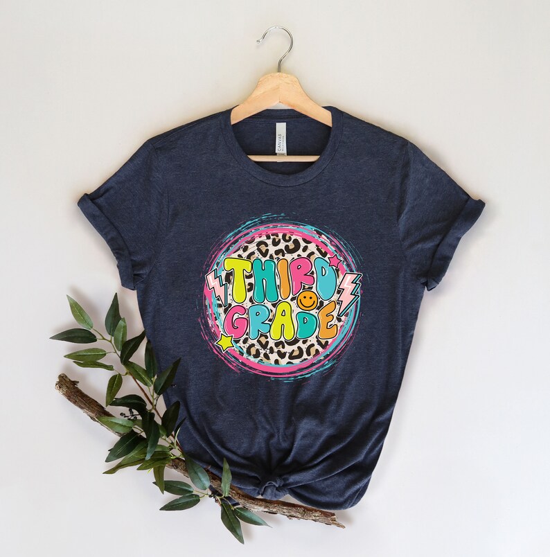 May include: A dark blue t-shirt with a colorful graphic design that says "Third Grade" in a retro font. The graphic features a leopard print circle with pink, blue, and yellow accents.