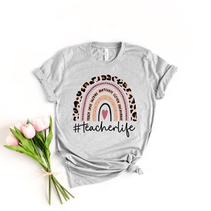 Puede incluir: Una camiseta gris con un dise&ntilde;o de arco&iacute;ris que incluye las palabras "teach, love, inspire, motivate, listen, empower" y un coraz&oacute;n rosa. El arco&iacute;ris est&aacute; bordeado con un estampado de leopardo. El texto "#teacherlife" est&aacute; impreso debajo del arco&iacute;ris.